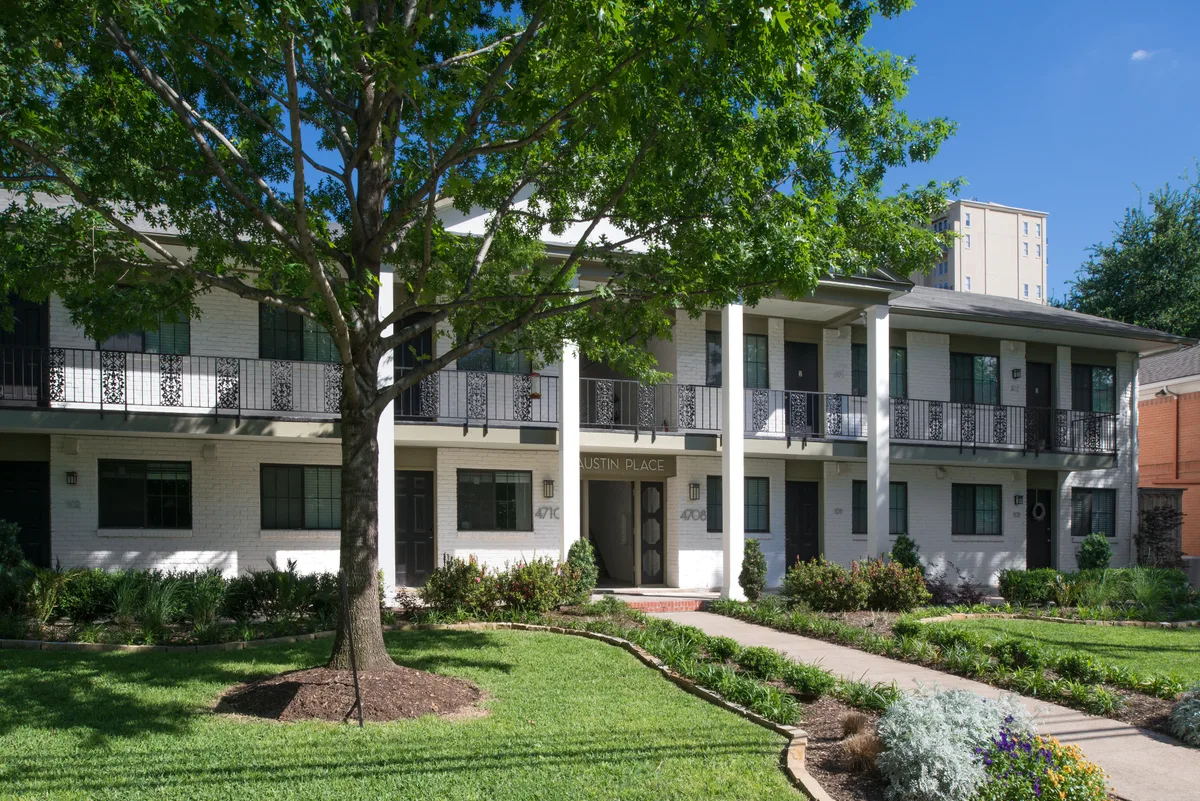 White brick two-story building framed by mature trees and flowering garden beds