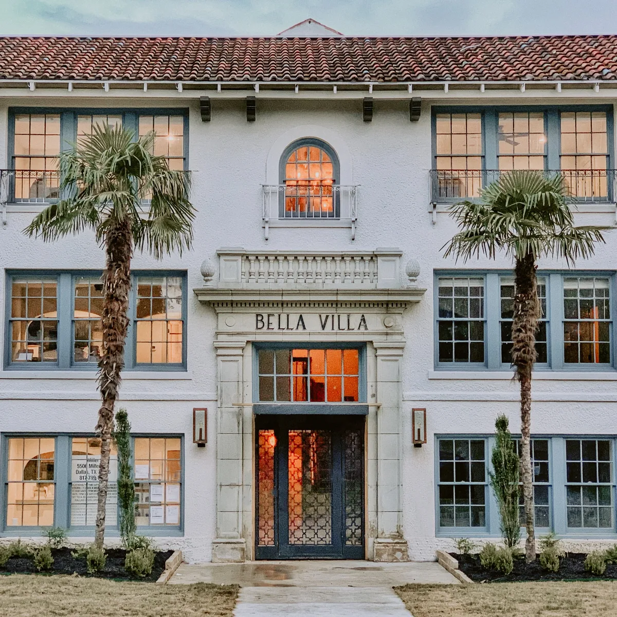 Ornate entry portico with BELLA VILLA stone lettering, arched window and glowing iron-and-glass doors between palms