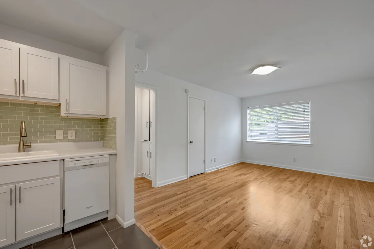 Sunlit living room with hardwood floors opening to a white kitchen with seafoam tile backsplash