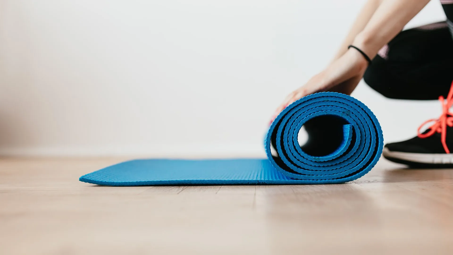 A rolled blue exercise mat on a wood floor beside a runner reaching down in athletic shoes