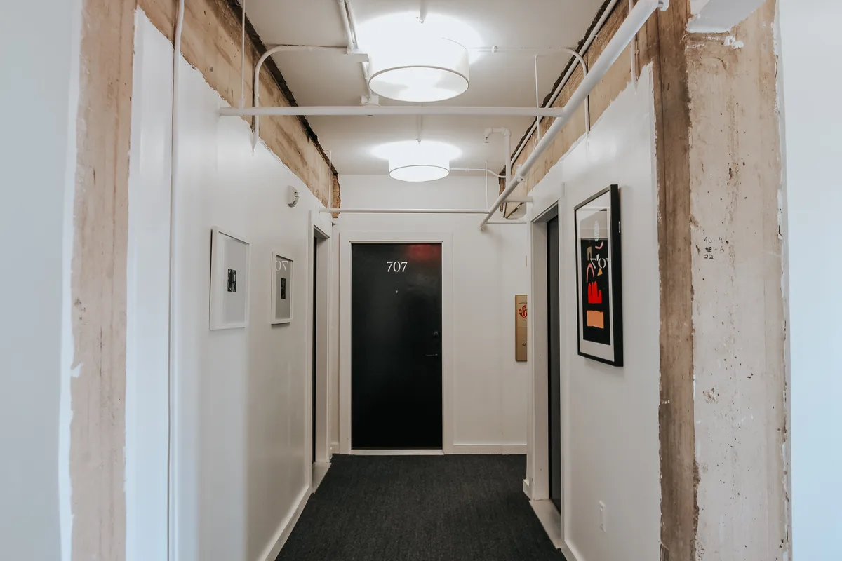 Residential hallway with exposed concrete columns, carpeted floor and framed art beside numbered unit doors