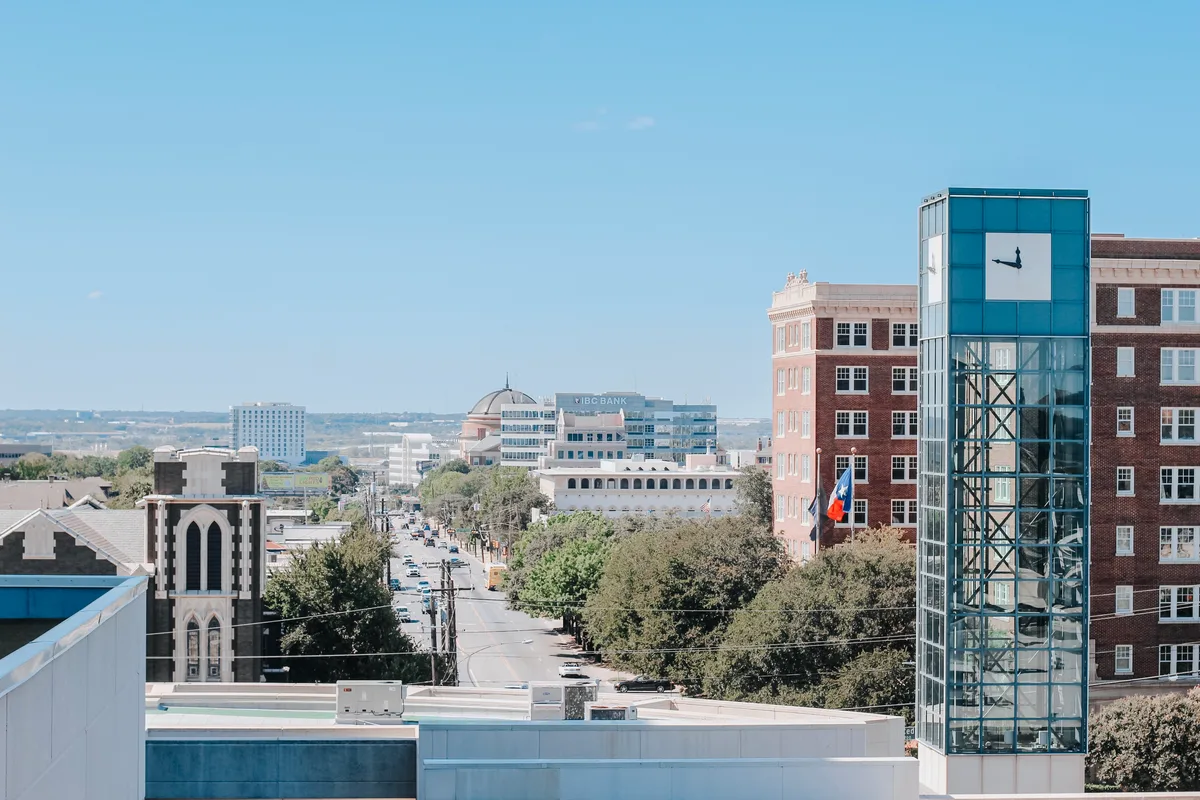 Elevated view over Oak Lawn rooftops toward the downtown Dallas skyline and a historic church steeple