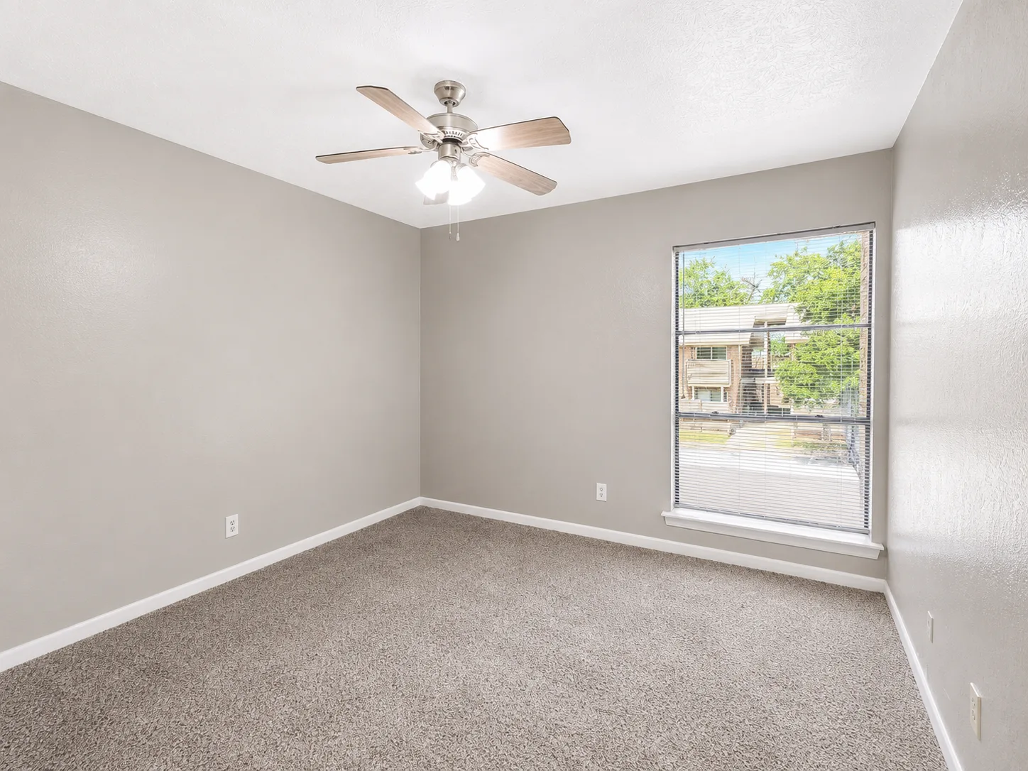Empty carpeted A1 bedroom with a ceiling fan and a blinded window looking out to a neighboring building