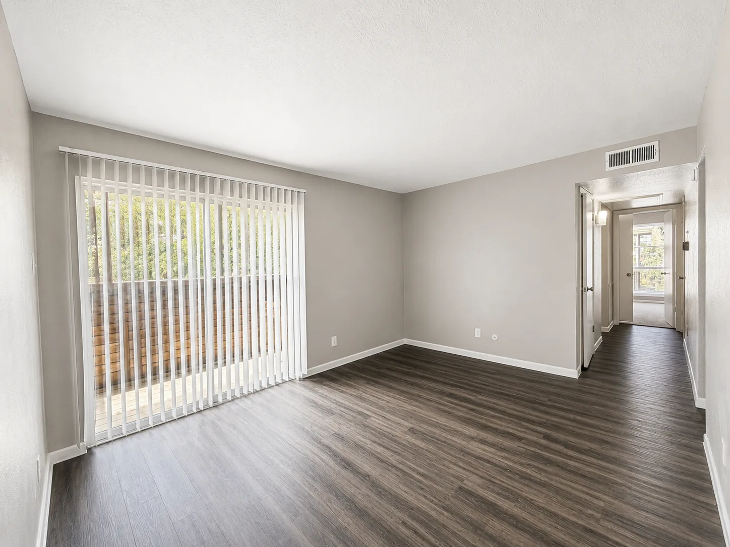 Empty living room in an A1 home with a sliding patio door behind vertical blinds, dark wood-look flooring, and a hallway leading toward the bedroom