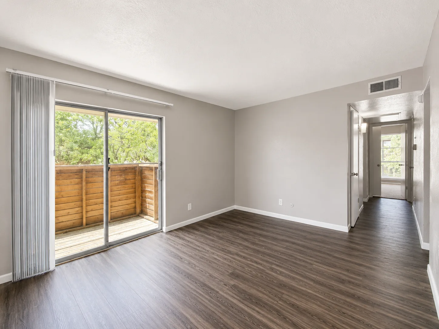 Empty A1 living room with a sliding glass door opening to a wood-fenced private patio, dark wood-look flooring, and a hallway beyond