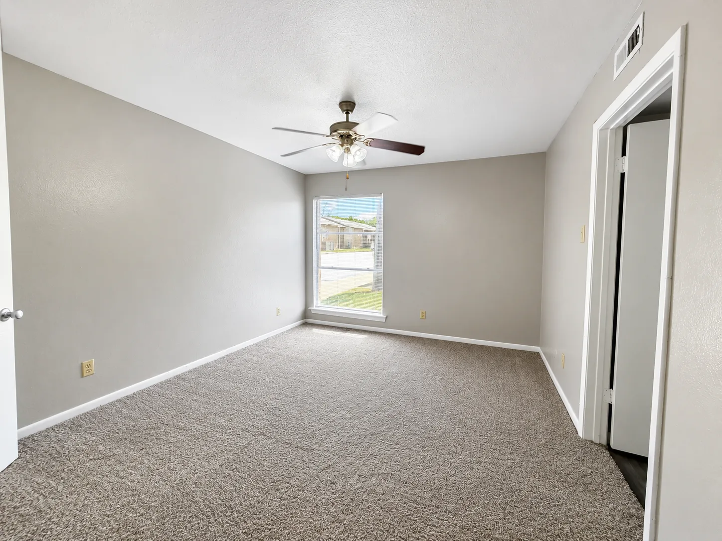Empty carpeted bedroom in a B1 home with a ceiling fan and a blinded window looking out to a single-story building