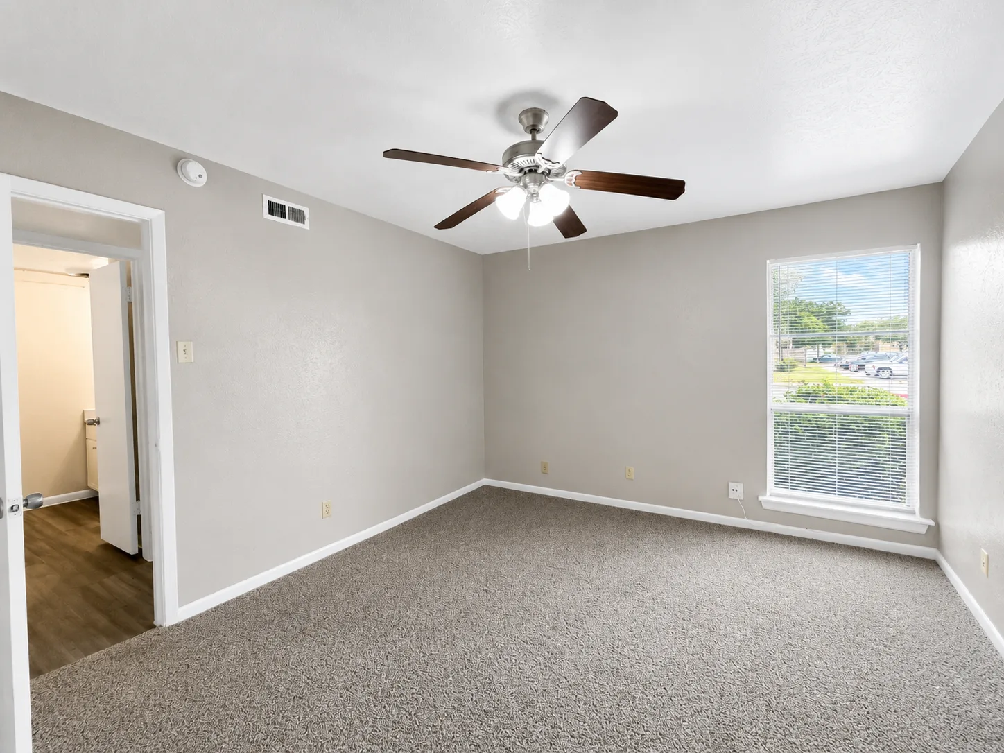 Empty carpeted B1 bedroom with a ceiling fan, a blinded window, and a doorway through to the bathroom