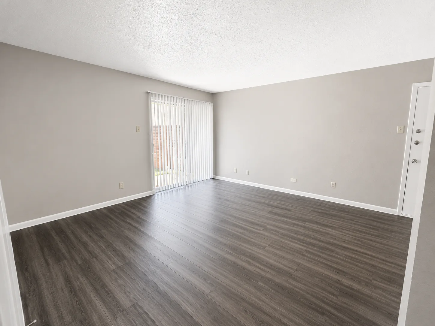 Empty B1 living room with a sliding patio door behind vertical blinds and wood-look flooring