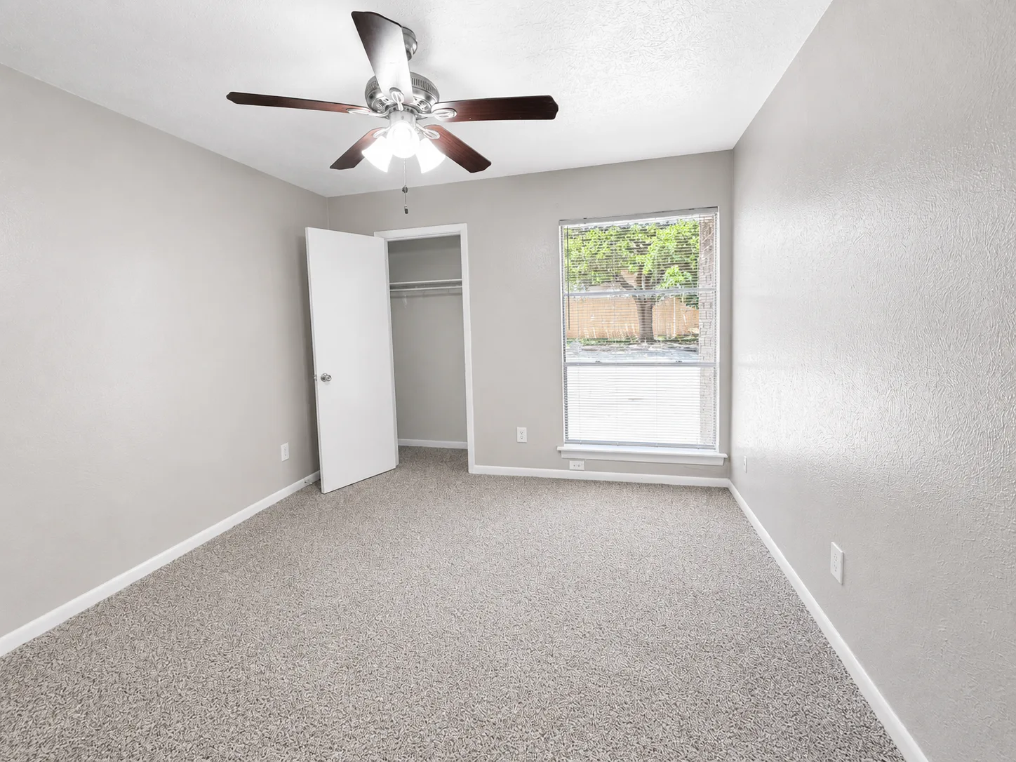Empty carpeted bedroom in a C1 home with a ceiling fan, an open closet with a hanging rod, and a window looking out to a tree and brick wall