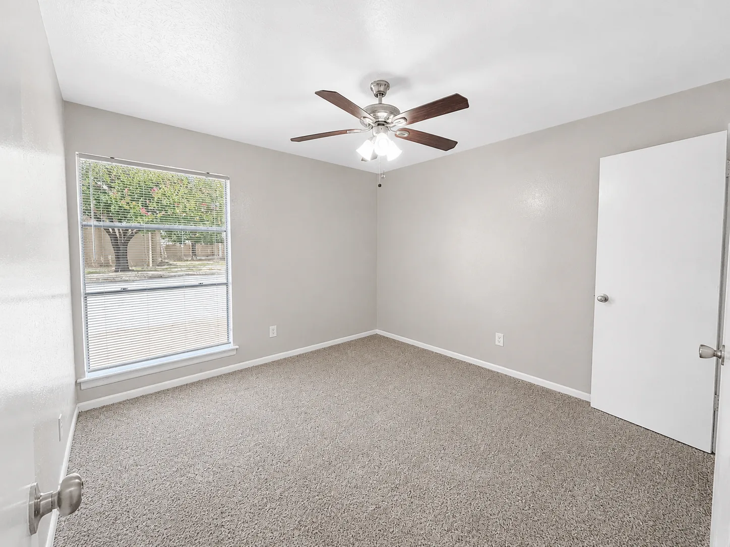 Empty carpeted C1 bedroom with a ceiling fan and a wide blinded window overlooking the grounds