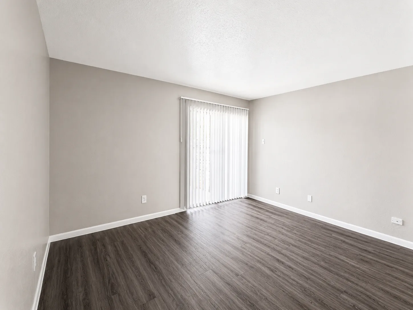 Empty C1 living room with a sliding patio door behind vertical blinds and wood-look flooring